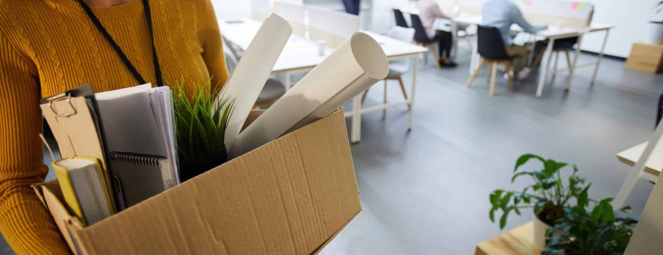 Serious sad woman in sweater carrying cardboard box full of stuff and leaving office after dismissal